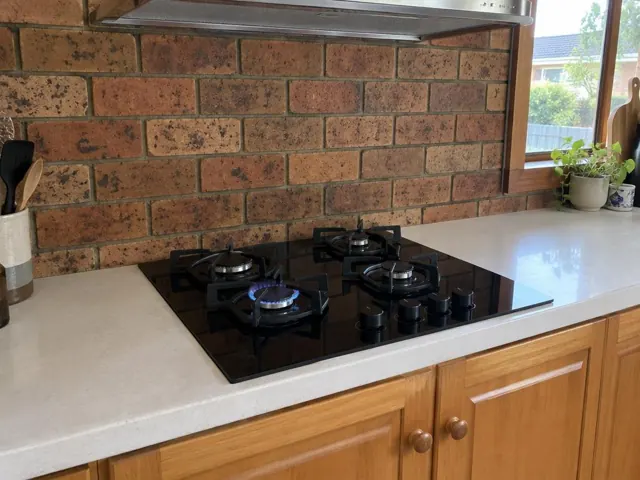 Black gas cooktop set into a light benchtop with brick splashback and timber cabinets in Cameron Park