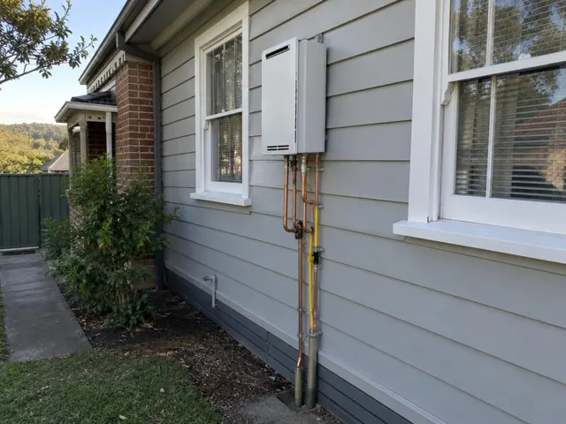 Wall-mounted hot water unit with copper and gas pipes beside windows on a weatherboard house in Minmi